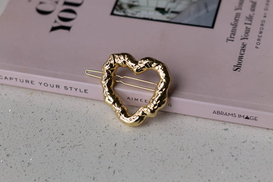 A gold metal irregular heart hairpin with a dented texture, styled on a pink hardcover fashion book.
