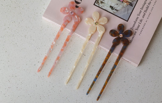 A collection of acetate hairpins with decorative flower-shaped toppers in the colour brown, pink, and white, with a little gem detail, displayed on a surface near a book.