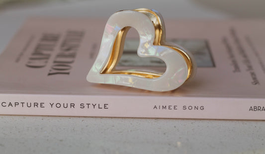 A white acetate heart-shaped hair claw placed on a book with the author's name, Aimee Song, visible.