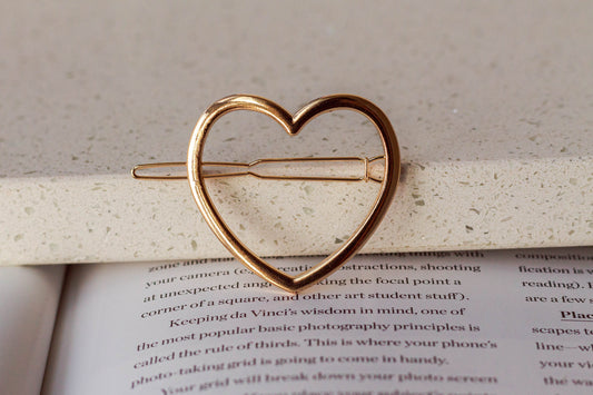 A metal heart hairpin in the colour gold is placed on a surface with a blurred background.