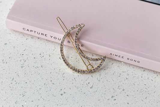 A gold-colored rhinestone moon-shaped hairpin placed on a pale pink book with the author's name, Aimee Song, inscribed on the cover.