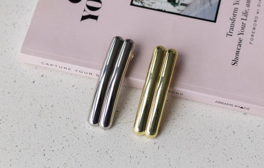 Silver and gold metal double-bar hair barrettes displayed side by side on a white speckled surface next to a pink hardcover book