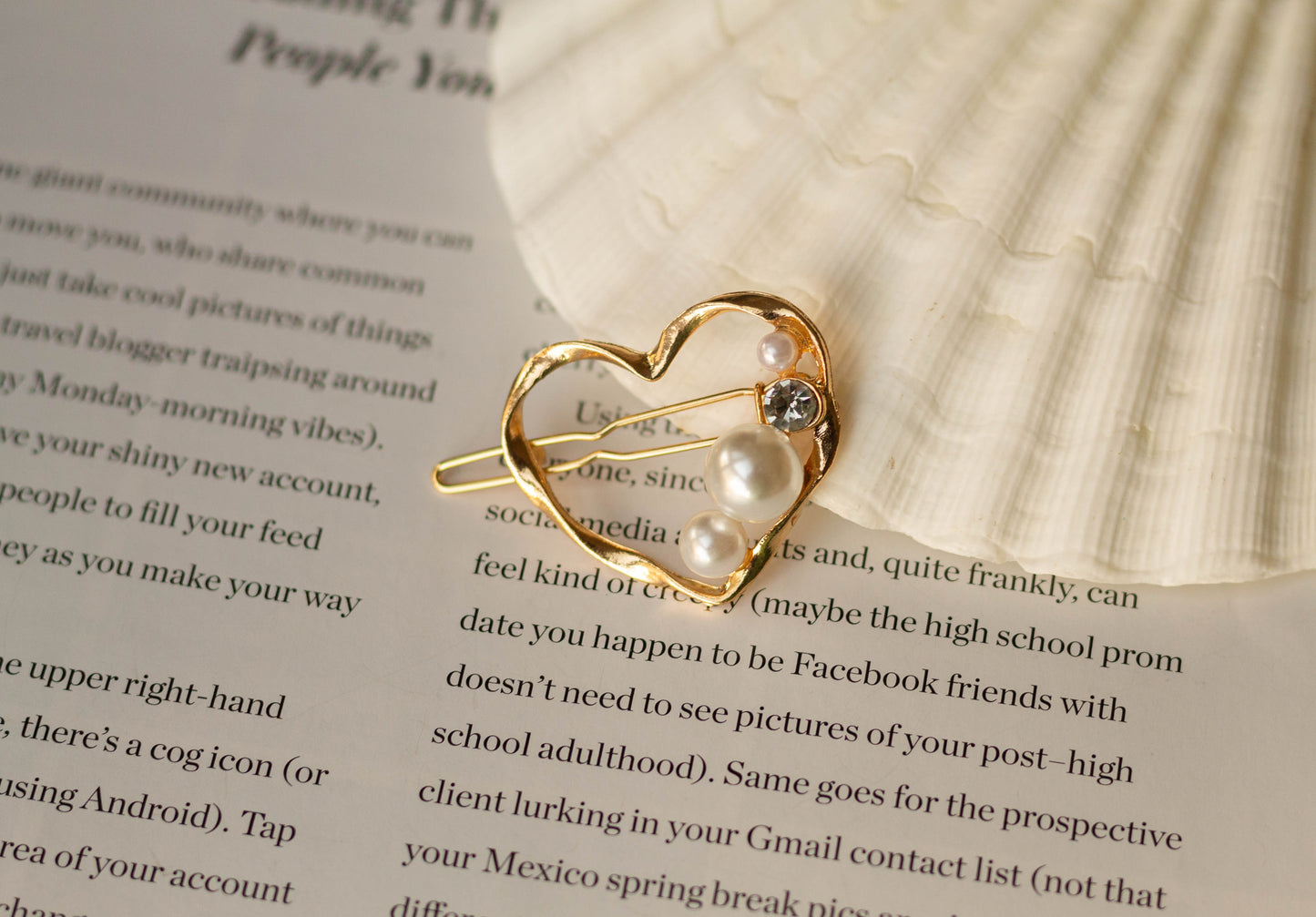Gold metal mini heart hairpin with pearl and gem accents, styled on a white seashell and an open book page.
