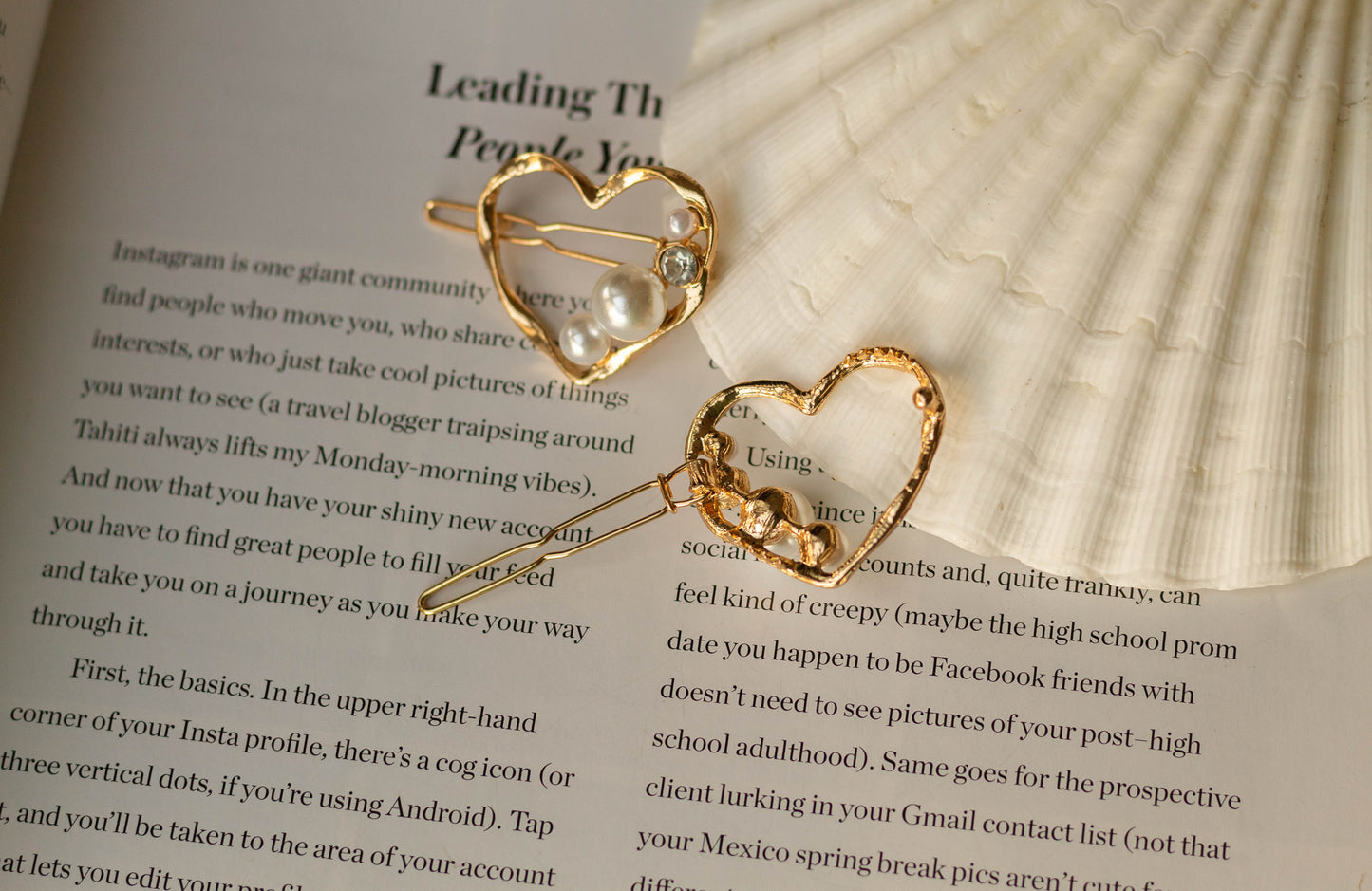 Back view of the gold metal mini heart hairpin with pearl and gem accents, showing clasp mechanism, styled on a white seashell and open book page.