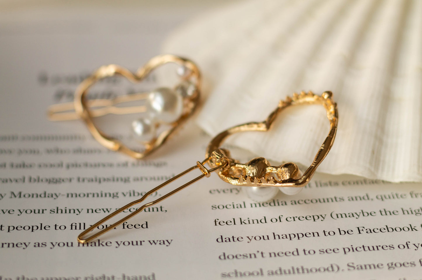 Back view of the gold metal mini heart hairpin with pearl and gem accents, showing clasp mechanism, styled on a white seashell and open book page.