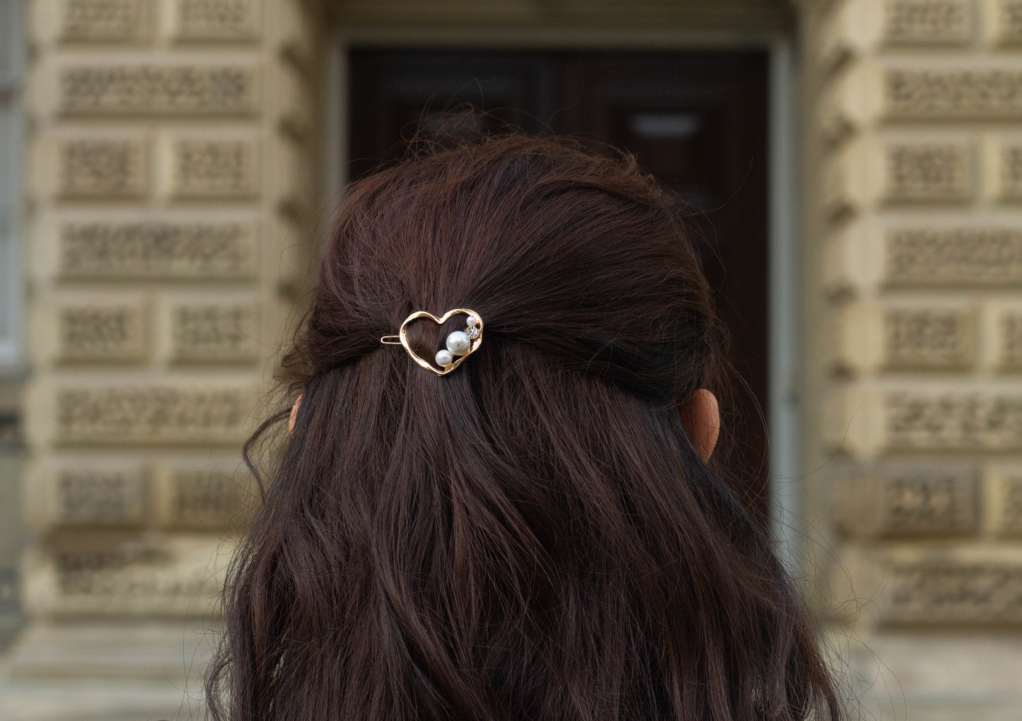 Back view of a woman with wavy dark brown hair wearing a gold metal heart hairpin adorned with pearl and gem accents, photographed in front of a historic stone building.