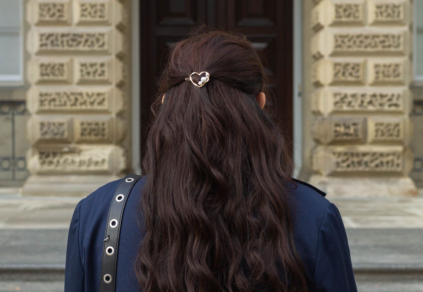 Back view of a woman with wavy dark brown hair wearing a gold metal heart hairpin adorned with pearl and gem accents, photographed in front of a historic stone building.