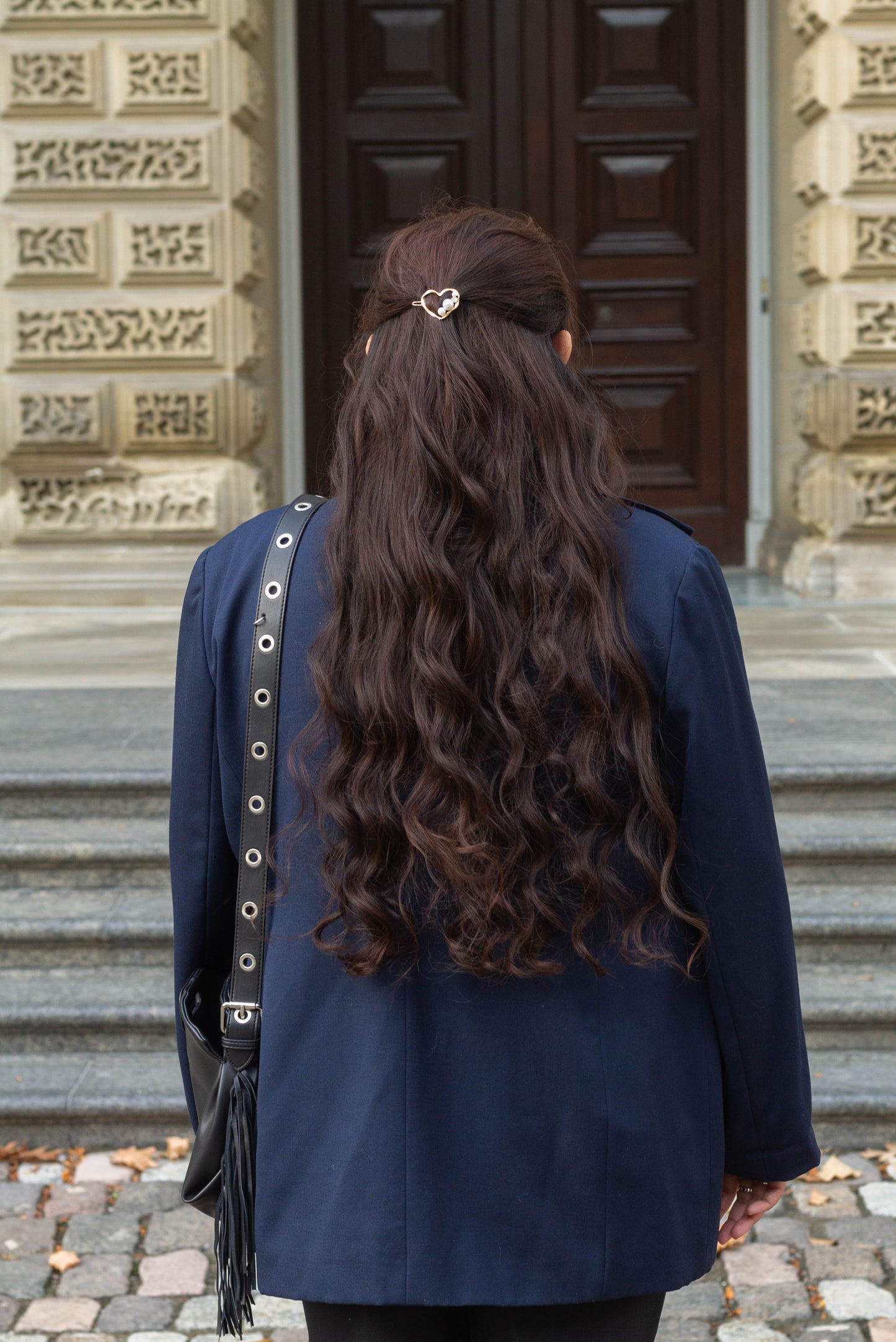 Back view of a woman with wavy dark brown hair wearing a gold metal heart hairpin adorned with pearl and gem accents, photographed in front of a historic stone building.