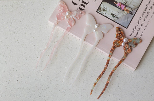 The french hairpins in the shape of a butterfly adorned with pearl details comes in the colours mix brown, mix pink, and white, resting on a pink fashion book.