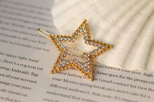 Close-up view of gold rhinestone star hairpin showing rhinestone details, styled on an open book with a seashell – celestial-inspired accessory for elegant hairstyles.