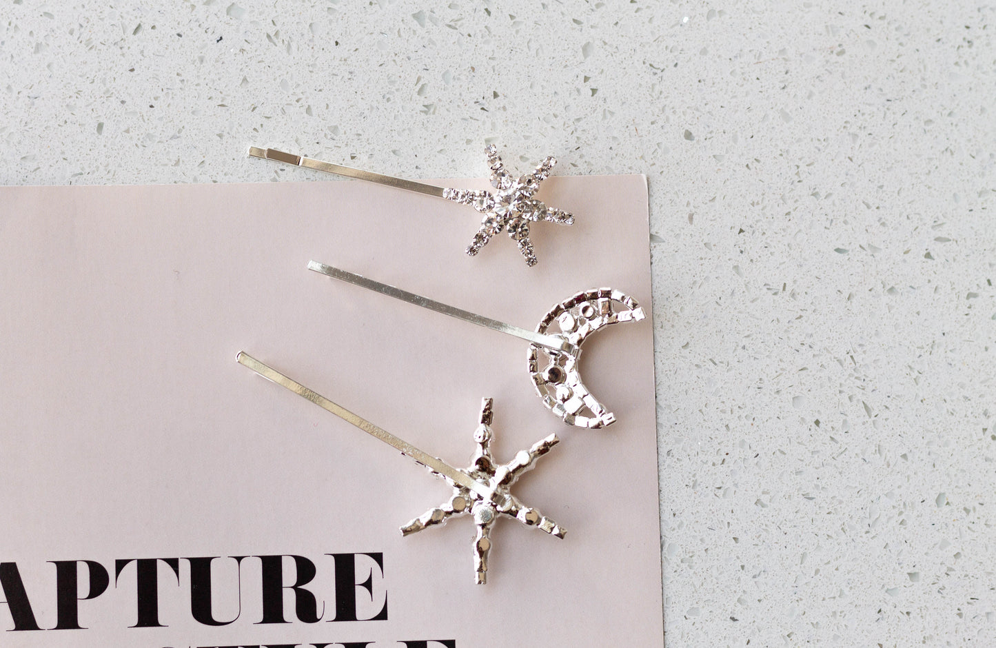 The rhinestone moon star hairpins are placed along a pink fashion magazine. The back side of the hairpins shows how they are soldered onto the hairpin.