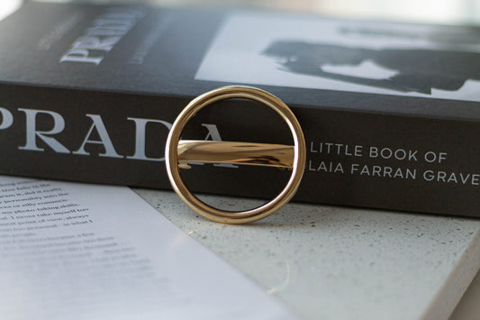 A gold-colored thick metal circle hair barrette placed on an open book with the title 'Prada' visible.