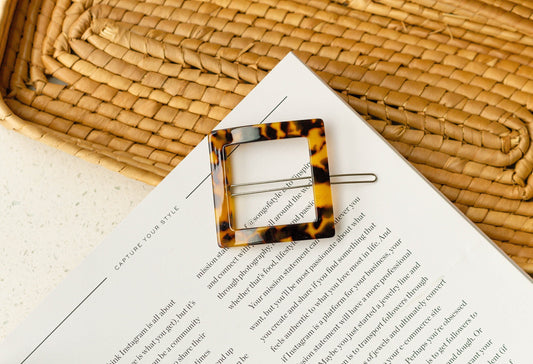 A tortoise shell square hairpin placed on top of a woven mat, with a blurred background of printed text.