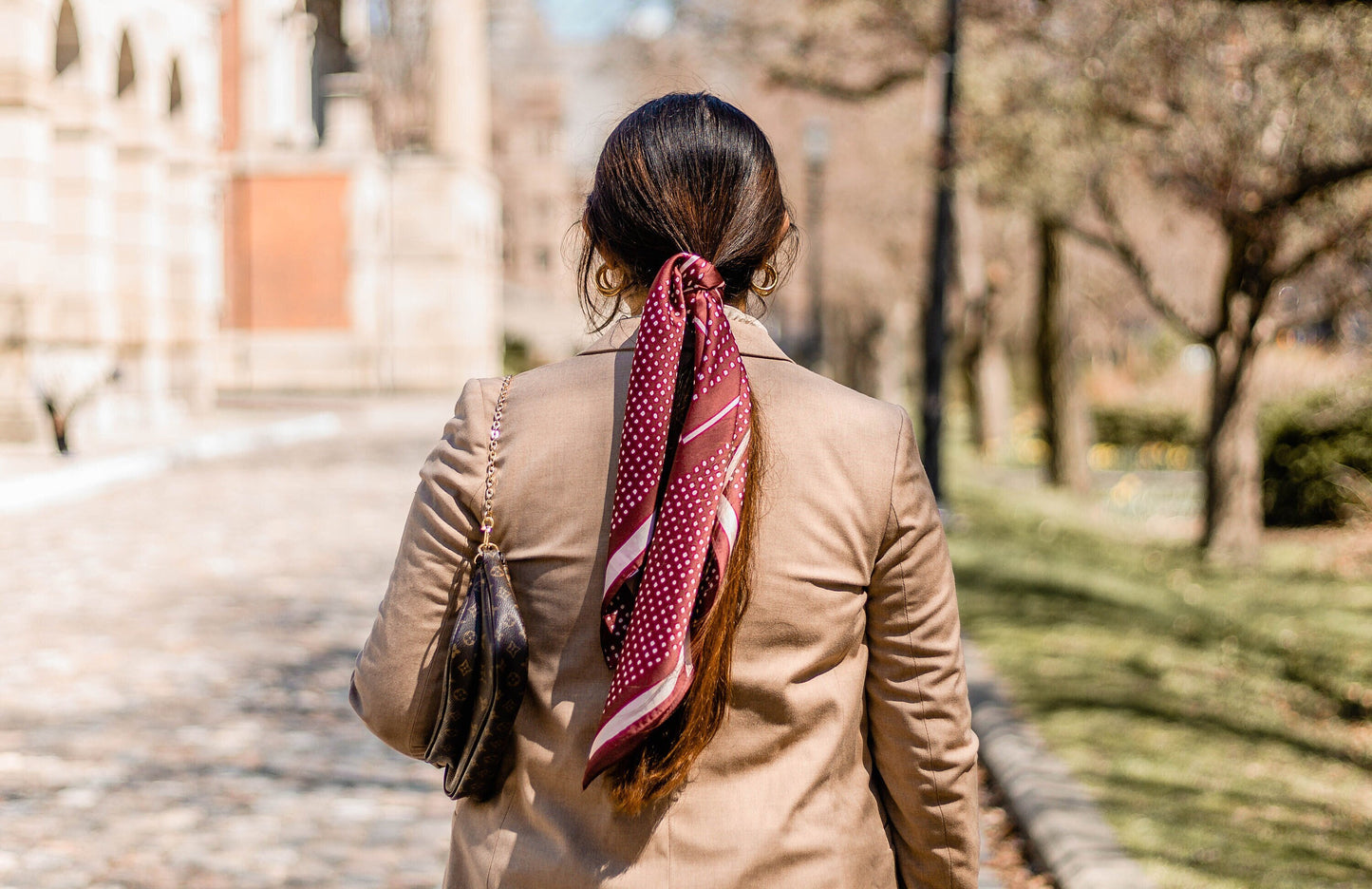 A model with long, straight brown hair is wearing a red hair scarf into a hair tie, standing in front of a historical building.