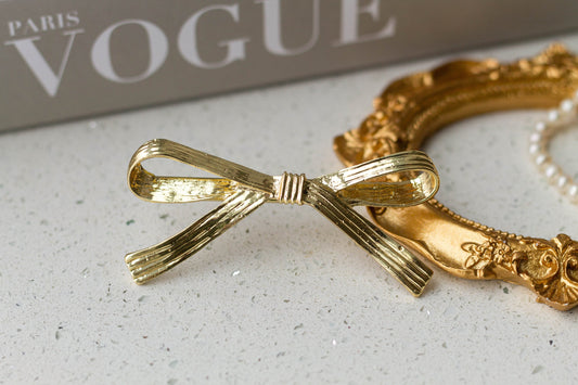 A gold-colored metal hair bow clip with an alligator-style fastener, placed on a surface next to a pearl necklace and a 'Paris Vogue' book.