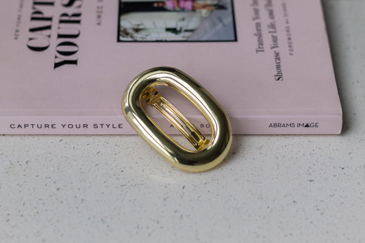 Close-up of gold metal oval barrette highlighting its shiny finish, displayed on a white speckled surface beside a pink hardcover book